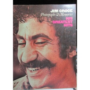 Puede incluir: Portada de álbum con un hombre sonriente con un gran bigote. La portada dice "JIM CROCE Photographs & Memories HIS GREATEST HITS" en texto blanco y rosa. La imagen tiene una apariencia vintage, ligeramente descolorida.