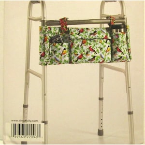 May include: A walker bag with a colorful bird and leaf pattern. The bag has multiple pockets for storage and is attached to a silver walker frame. A black travel mug and a small electronic device are visible.