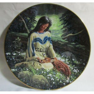 Puede incluir: Un plato de porcelana con una pintura de una mujer nativa americana con un vestido blanco y azul, sentada en un bosque con un cervatillo. La mujer está sosteniendo el cervatillo en sus brazos y lo está mirando. El fondo es un bosque verde exuberante con árboles y flores.