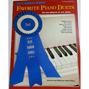Puede incluir: Una cinta azul con estrellas doradas y las palabras "BLUE RIBBON SERIES" en ella. La cinta está atada alrededor de un teclado de piano. La portada del libro dice "FAVORITE PIANO DUETS" y "Nivel Dos (Volumen Uno)".