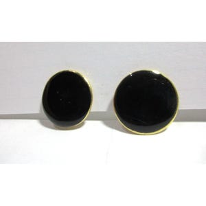 Puede incluir: Dos pendientes redondos negros con borde dorado. Los pendientes son circulares y tienen un acabado brillante. Se presentan sobre un fondo blanco.