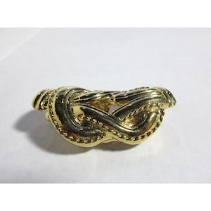 Puede incluir: Un nudo decorativo de metal dorado, con un diseño detallado e intrincado. El nudo es un elemento de diseño popular en joyería y decoración del hogar.