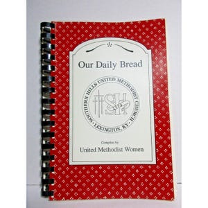 Puede incluir: Un libro rojo encuadernado en espiral con una cubierta blanca. La cubierta tiene un título que dice "Nuestro Pan Diario" y un logotipo para la Iglesia Metodista Unida Southern Hills en Lexington, Kentucky. El texto "Compilado por las Mujeres Metodistas Unidas" está impreso debajo del logotipo.