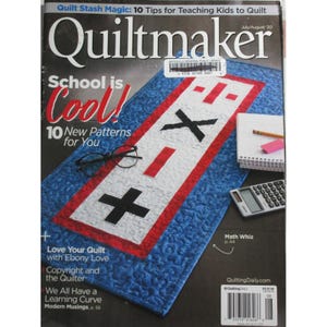 May include: The cover of a "Quiltmaker" magazine features a blue quilt with a red and white design of math symbols. The text "School is Cool! 10 New Patterns for You" is displayed. Other items include glasses, a notepad, and a calculator.