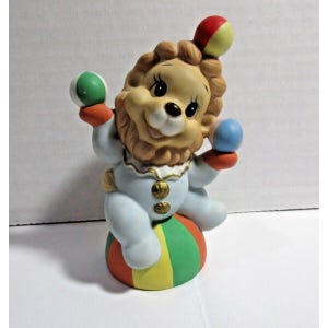 May include: A porcelain figurine of a lion wearing a clown costume and balancing on a ball. The lion is holding three colorful balls in its paws. The figurine is painted in bright colors, including blue, red, green, and yellow.