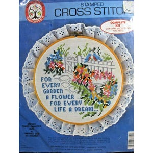 May include: A stamped cross stitch kit featuring a colorful floral design with a white picket fence and a birdhouse. The kit includes everything you need to complete the project, including a 10-inch hoop, embroidery floss, needle, and instructions. The kit is titled "For Every Garden A Flower For Every Life A Dream."