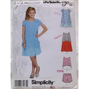 Puede incluir: Un patrón de costura Simplicity para ropa de niña. El patrón presenta a una niña pequeña con un vestido azul claro con detalles de encaje. El patrón también muestra ilustraciones de otros conjuntos, incluyendo un top a rayas y una falda roja, y pantalones cortos estampados.