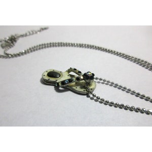 Pode incluir: Colar prateado com um pendente em forma de chinelo. O pendente tem uma base creme com detalhes pretos e iridescentes. Uma pequena flor adorna a correia. A corrente é feita de pequenas contas redondas. Um acessório com tema de verão.