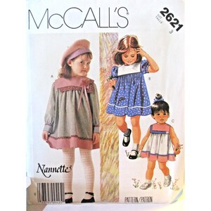Puede incluir: Patrón de costura McCall's para vestidos de niña, talla 3. El patrón presenta tres diseños de vestidos, incluyendo un vestido de manga larga con cuello y ribete rosa, un vestido floral azul y un vestido sin mangas con detalles en rosa. El patrón incluye el logo "Nannette".