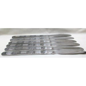 May include: A set of seven stainless steel dinner knives. The handles feature a decorative, repeating pattern of squares with ornate designs. The blades are smooth and curved, reflecting the light. The knives are arranged in a fan shape.