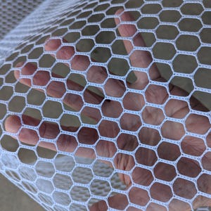 May include: Close-up of a white hexagonal mesh fabric. The mesh is held up, revealing the texture and pattern of the material. The fabric is a bright white color.