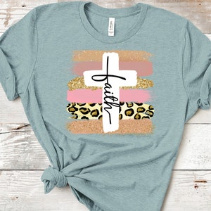 May include: A light blue t-shirt with a white cross and the word "faith" written on it. The cross is surrounded by pink and gold brushstrokes and a leopard print pattern.