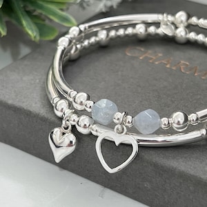 May include: Silver bracelets featuring heart charms and pale blue gemstone beads. The bracelets are made of polished metal and have a delicate design. The charms are in the shape of a solid heart and a heart outline.