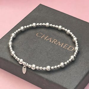 May include: A stretch bracelet composed of polished silver-toned beads of varying sizes arranged in a repeating pattern. A small, oval-shaped silver-toned charm hangs from the bracelet, featuring the engraved number 925 on its surface.