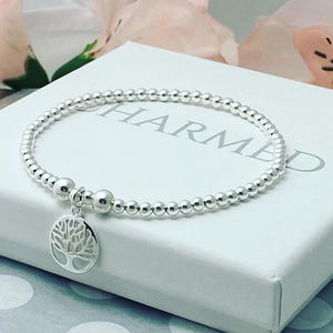 May include: A silver bracelet with small, round beads and a tree of life charm. The charm is a detailed tree design. The bracelet is presented on a white box with the word "CHARMED" printed on it. The bracelet is a delicate piece of jewellery.
