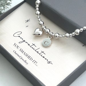 May include: A silver beaded bracelet with a heart charm and a round charm with the letter "C". The bracelet is presented in a grey box with the words "Congratulations YOU SMASHED IT!" on a white card.
