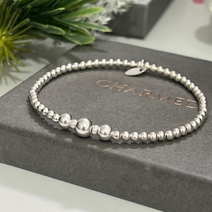 May include: A silver beaded bracelet with three larger beads at the centre. The bracelet is presented on a grey box with the word "CHARMED" embossed on the lid. The bracelet is made of small, round beads.