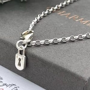May include: A silver-toned chain necklace featuring a small padlock charm with a keyhole cutout. The necklace includes a lobster claw clasp and is displayed on a dark grey box with the partial text HARMED visible in copper-coloured lettering.