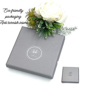 May include: A square grey gift box featuring a circular logo with a lotus flower and the text CHARMED. The box is adorned with a white rose and green foliage. Text on the image reads Eco friendly packaging and Anti tarnish insert.