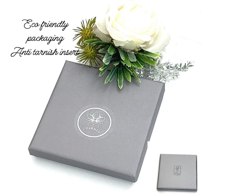 May include: A square grey gift box featuring a circular logo with a lotus flower and the text CHARMED. The box is adorned with a white rose and green foliage. Text on the image reads Eco friendly packaging and Anti tarnish insert.
