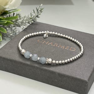May include: Silver beaded bracelet featuring three pale blue gemstones. The bracelet rests on a grey box with the word "CHARMED" printed on it. The bracelet has a small charm. Jewellery.