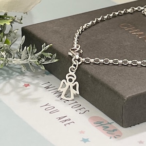 May include: A silver bracelet featuring an angel charm. The bracelet is presented on a dark grey box, with the words "Twinkle Toes You Are" printed on a white card. The background includes a sprig of greenery.