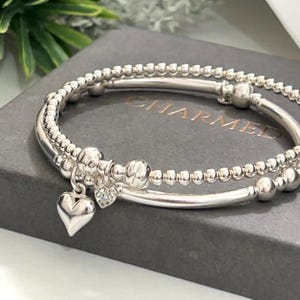 May include: Two silver bracelets with a heart charm. The bracelets are made of small beads and are stacked on top of each other. The heart charm is attached to the bottom bracelet.