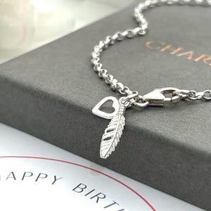 May include: A silver chain bracelet featuring a small open heart charm and a textured feather charm. The bracelet is secured with a lobster clasp and rests on a dark grey box with the word CHARM visible, near a card with the text HAPPY BIRTHDAY.