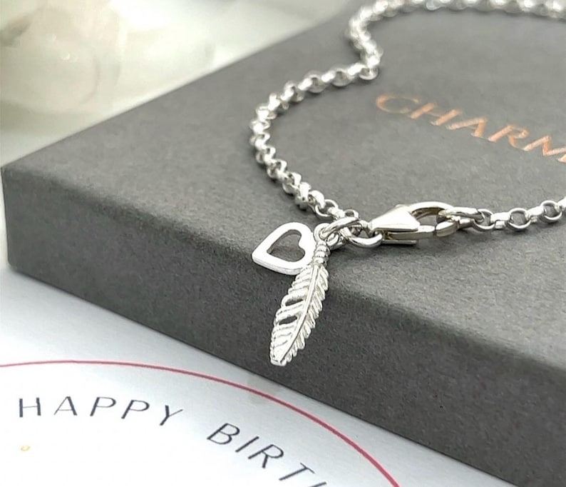 May include: A silver chain bracelet featuring a small open heart charm and a textured feather charm. The bracelet is secured with a lobster clasp and rests on a dark grey box with the word CHARM visible, near a card with the text HAPPY BIRTHDAY.