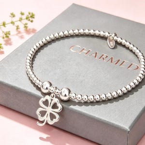 May include: A silver beaded stretch bracelet featuring a four-leaf clover charm and two larger silver beads. A small oval tag marked 925 is attached to the bracelet. The jewellery is displayed on a grey box with the word CHARMED printed in rose gold.