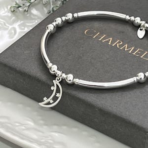 May include: A silver-toned stretch bracelet featuring alternating round beads and smooth cylindrical segments. A crescent moon charm with small star cutouts hangs from the bracelet. The jewellery is displayed on a dark grey box with the text CHARMEL.