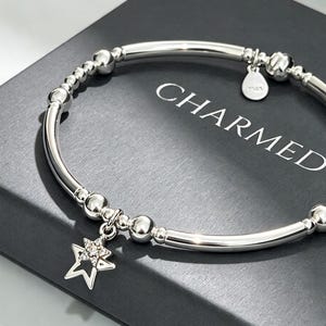 May include: A silver-toned stretch bracelet featuring alternating smooth round beads and elongated tube beads. A star-shaped charm with small embedded stones hangs from the bracelet, alongside a small circular tag with the text CHARMED.