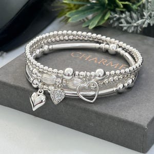 May include: A collection of silver bracelets, each with a different design and heart-shaped charms. The bracelets feature beaded and smooth sections. One charm is a solid heart, another is gem-studded, and the third is an open heart. The word "CHARME" is visible.