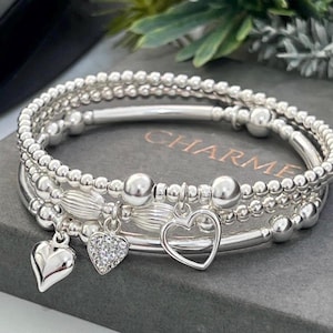 May include: A stack of three silver-toned beaded bracelets featuring a smooth metal tube, round beads, and three charms: a solid heart, a crystal-encrusted heart, and an open heart outline. The word CHARME is visible in the background.