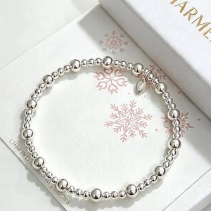 May include: A stretch bracelet composed of polished silver-toned spherical beads of varying sizes, arranged in a repeating pattern. A small, smooth, leaf-shaped silver-toned charm hangs from the bracelet, which rests on a white box with snowflakes.