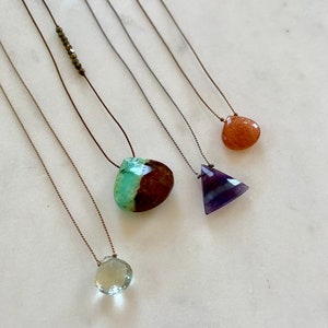 May include: Five necklaces with different colored gemstones. The necklaces are made with brown string and feature a green and brown stone, a purple triangle stone, a light brown stone, and a light green stone.