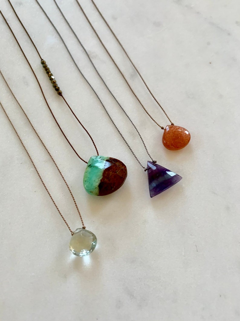May include: Five necklaces with different colored gemstones. The necklaces are made with brown string and feature a green and brown stone, a purple triangle stone, a light brown stone, and a light green stone.