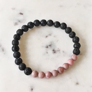 Puede incluir: Un brazalete de cuentas de piedra de lava negra y piedras preciosas rosas. El brazalete está hecho con pequeñas cuentas redondas.