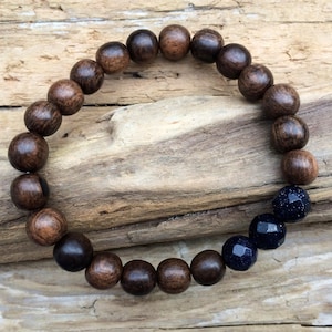 Puede incluir: Un brazalete de cuentas de madera marrón oscuro con un acento de piedra preciosa azul oscuro. El brazalete está hecho de cuentas de madera redondas y una sola piedra preciosa azul oscuro facetada.