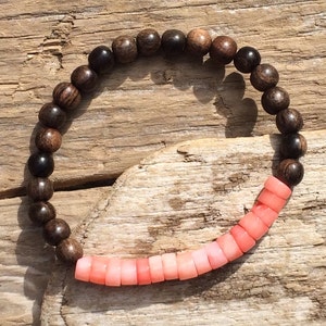 Puede incluir: Una pulsera con cuentas de madera marrón oscuro y cuentas cilíndricas de color coral. Las cuentas de madera son redondas y tienen un patrón de vetas de madera natural. Las cuentas de coral varían de rosa claro a oscuro.