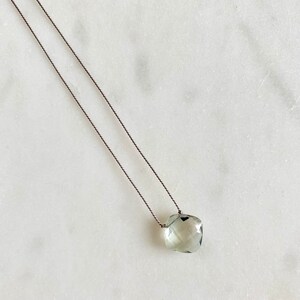 May include: A delicate necklace with a light green gemstone pendant. The pendant is faceted and has a slightly irregular shape. The necklace is made with a thin, dark grey cord.