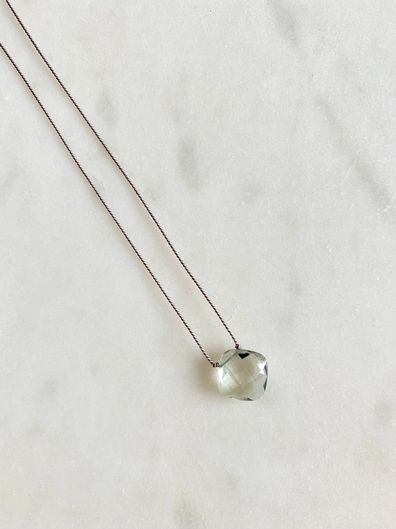 May include: A delicate necklace with a light green gemstone pendant. The pendant is faceted and has a slightly irregular shape. The necklace is made with a thin, dark grey cord.