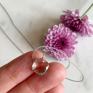 May include: A delicate silver necklace with a clear, faceted gemstone pendant. The pendant is a square shape with rounded edges and is suspended from a thin silver chain.