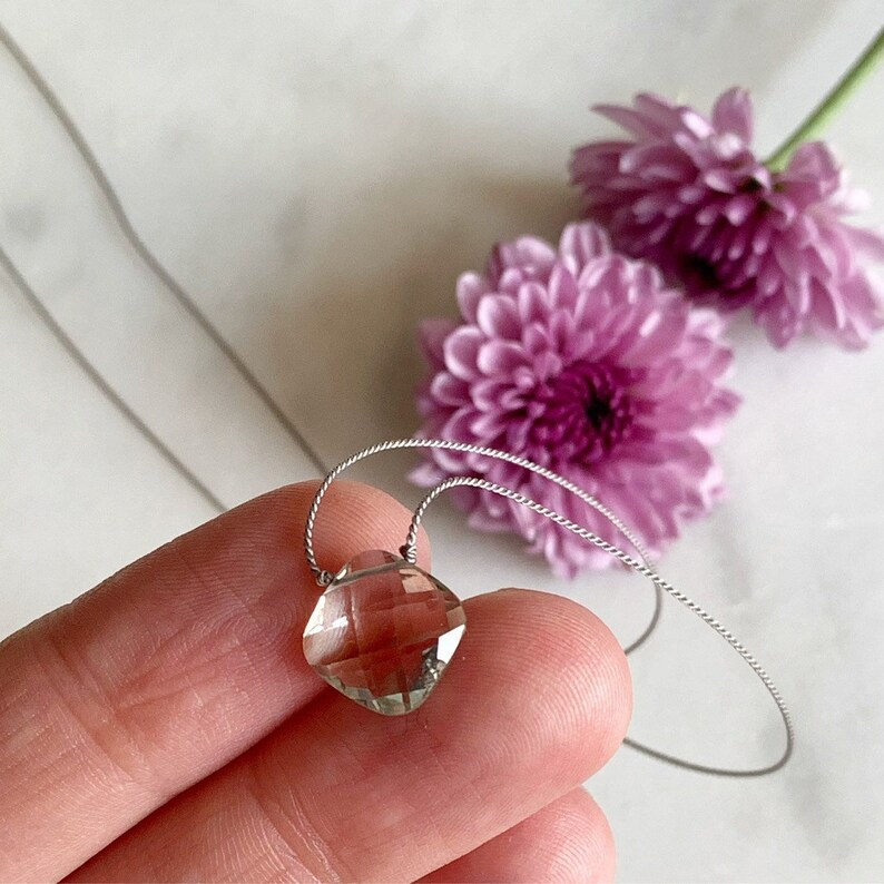 May include: A delicate silver necklace with a clear, faceted gemstone pendant. The pendant is a square shape with rounded edges and is suspended from a thin silver chain.