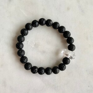 Könnte beinhalten: Ein Armband aus schwarzen Lavasteinen mit einem Anhänger aus klarem Quarzkristall.