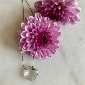 May include: A delicate necklace with a light green faceted gemstone pendant hanging from a thin brown cord. The necklace is displayed on a white surface with two pink flowers.