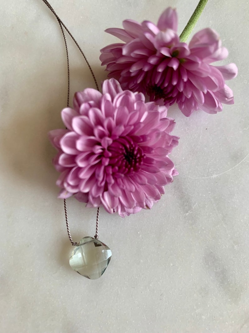 May include: A delicate necklace with a light green faceted gemstone pendant hanging from a thin brown cord. The necklace is displayed on a white surface with two pink flowers.