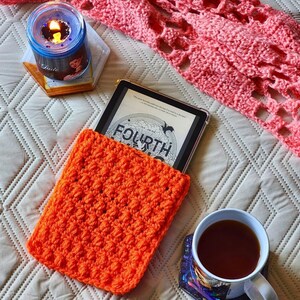May include: Orange crocheted book cover with a black and white book titled "Fourth" inside. The book cover is on a white surface with a pink crocheted blanket and a candle in the background.