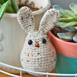 May include: A crocheted bunny toy with brown speckles, black eyes, and an orange nose. The bunny is sitting on a white shelf with a green plant in a white pot behind it.