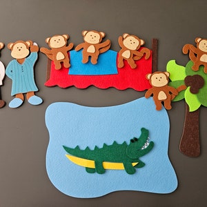 May include: Felt toy set featuring a brown alligator, a brown tree with green leaves, and six brown monkeys. Two of the monkeys are on a red bed with blue and white bedding. Two brown figures with white and blue clothing stand to the left of the bed.